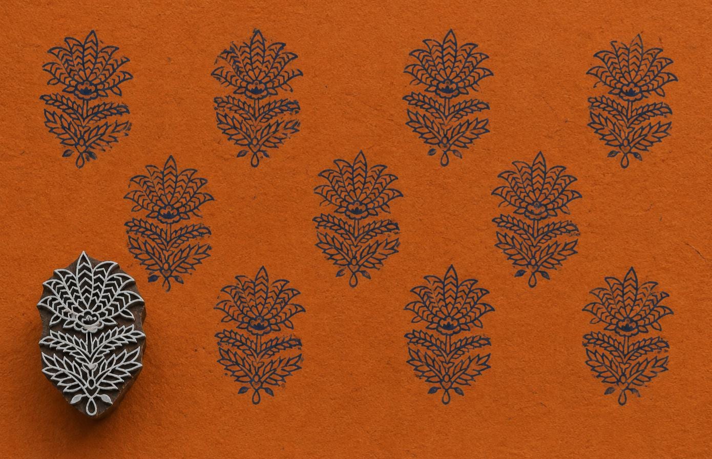 Hand-carved Indian floral wooden printing block displayed beside repeated dark blue botanical impressions on textured orange handmade paper.