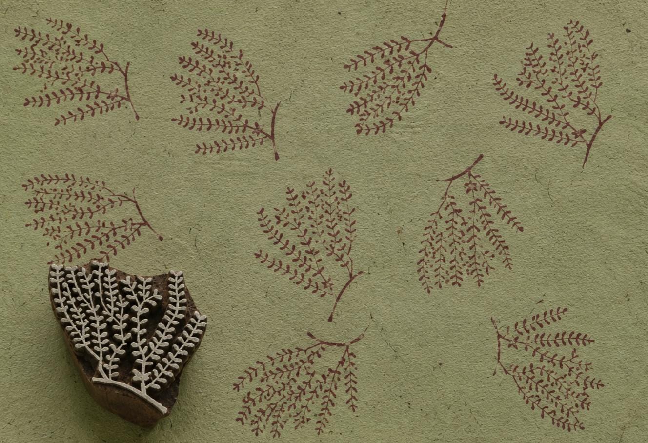 Hand-carved fern botanical wooden printing block beside repeated burgundy leafy branch impressions on textured sage green handmade paper.