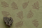 Hand-carved fern botanical wooden printing block beside repeated burgundy leafy branch impressions on textured sage green handmade paper.