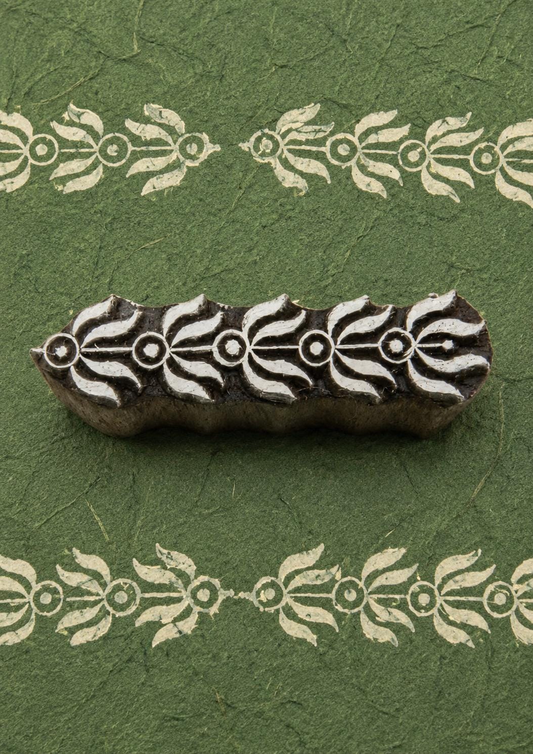 Hand-carved floral vine border wooden printing block with repeated cream leafy border impressions on textured dark green handmade paper.