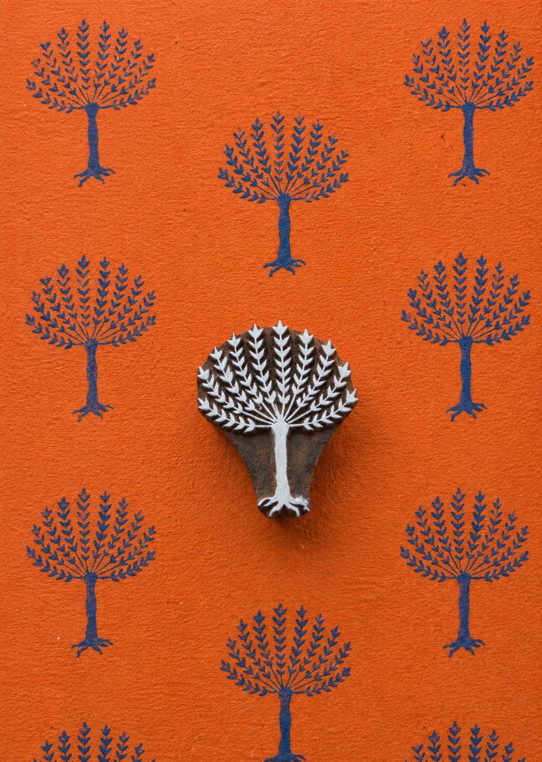 Hand-carved leafy tree wooden printing block surrounded by repeated dark blue tree impressions on textured orange handmade paper.