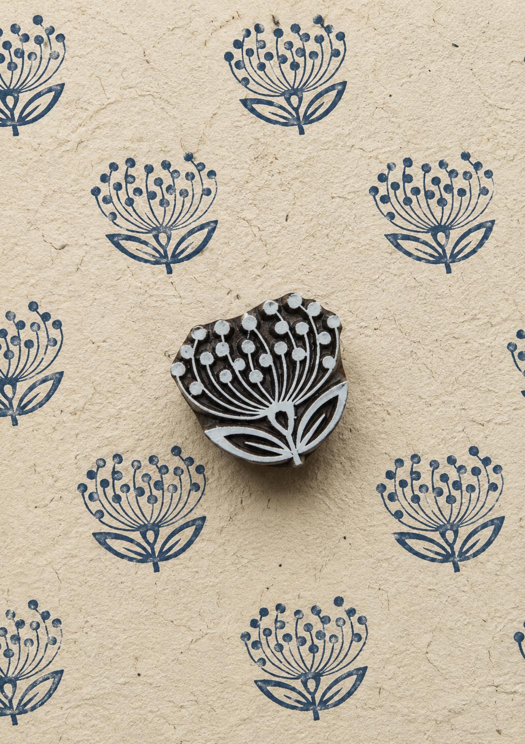Hand-carved dandelion flower wooden printing block surrounded by repeated navy blue botanical impressions on textured handmade paper.