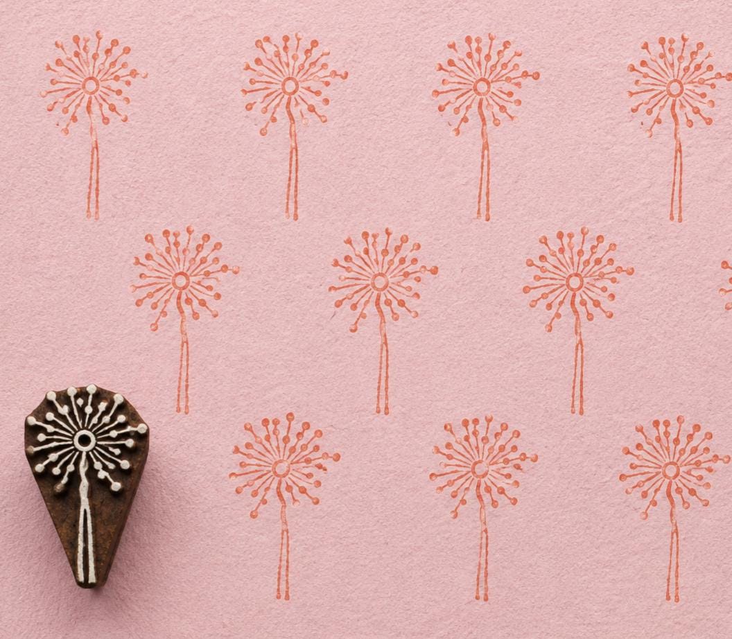 Hand-carved dandelion wooden printing block with repeated coral dandelion impressions on textured pale pink handmade paper.