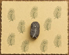 Hand-carved peacock feather wooden printing block surrounded by repeated olive green feather impressions on warm yellow handmade paper.