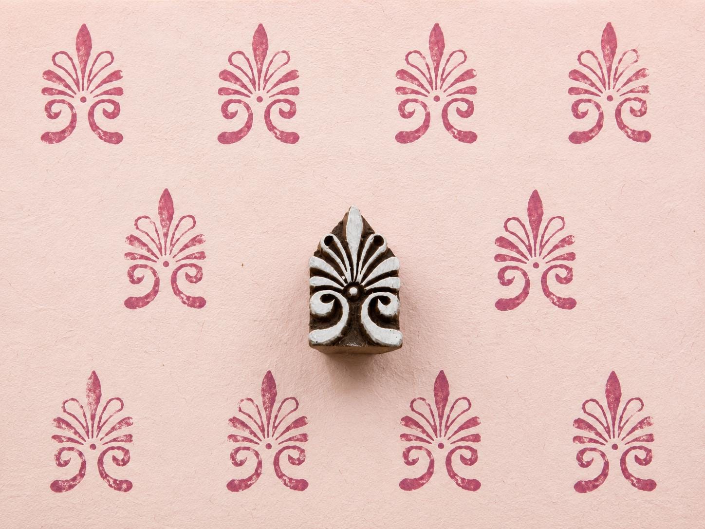 Hand-carved ornamental fleur-de-lis wooden printing block surrounded by repeated raspberry pink impressions on pale pink textured handmade paper.