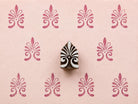 Hand-carved ornamental fleur-de-lis wooden printing block surrounded by repeated raspberry pink impressions on pale pink textured handmade paper.