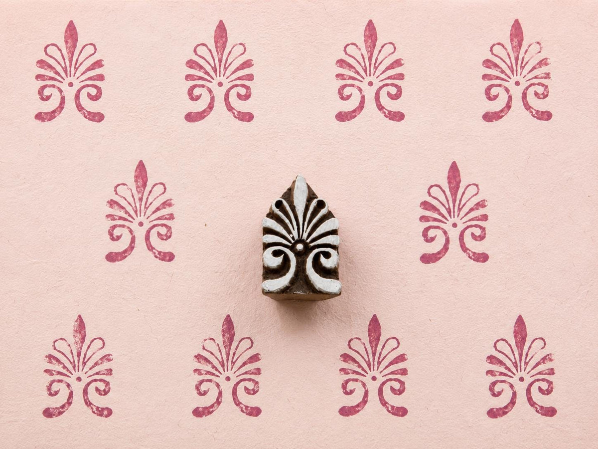 Hand-carved ornamental fleur-de-lis wooden printing block surrounded by repeated raspberry pink impressions on pale pink textured handmade paper.