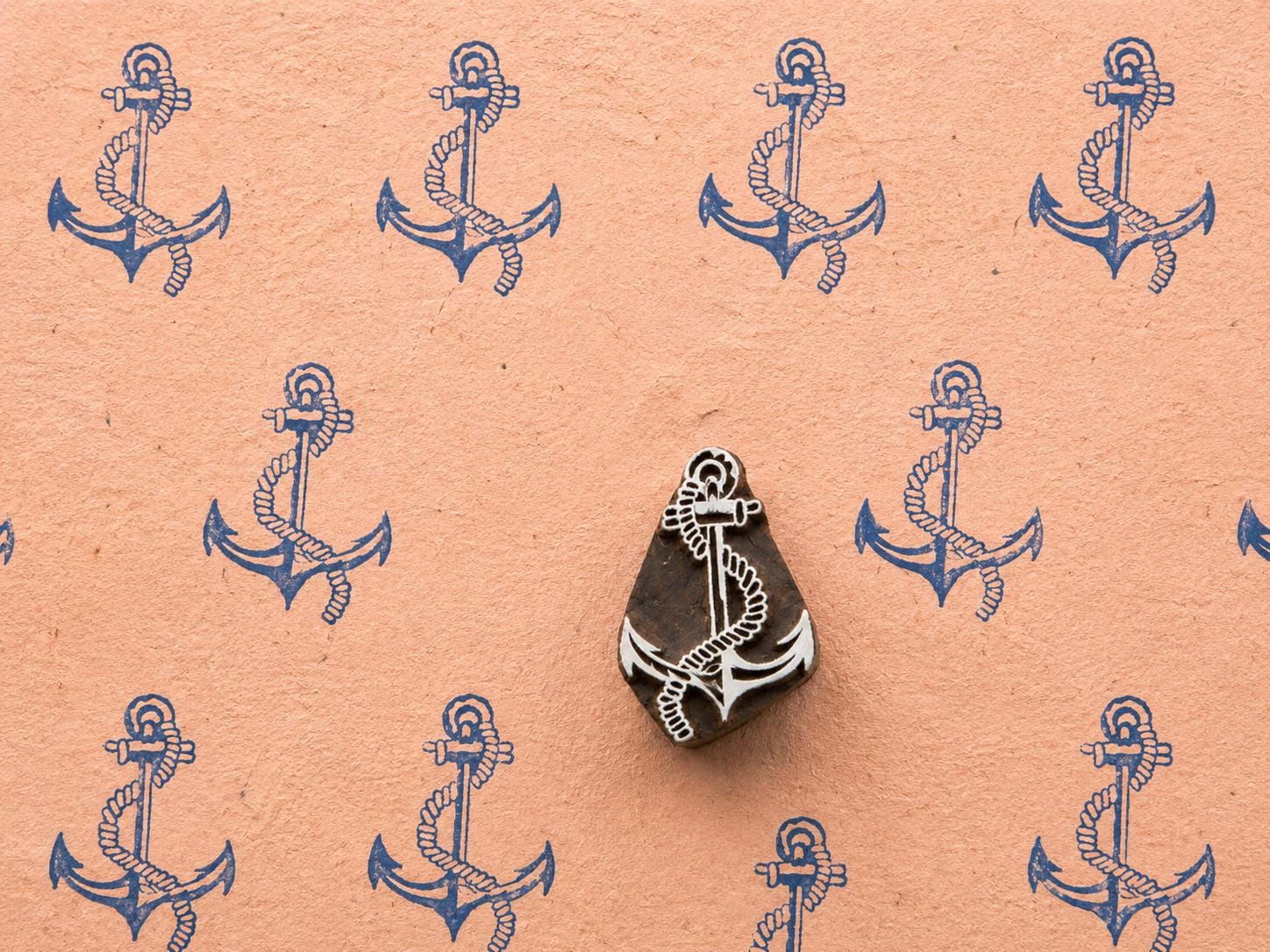 Hand-carved wooden anchor printing block with rope detail surrounded by repeated blue anchor prints on textured peach handmade paper.