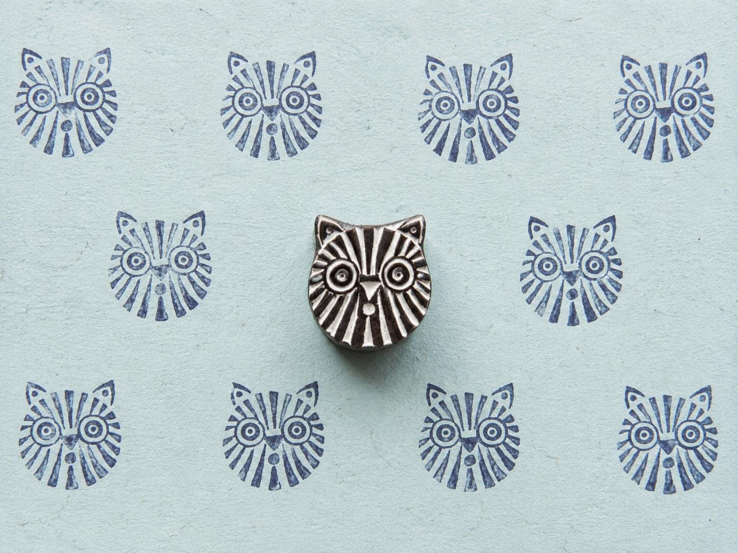 Hand-carved tribal cat wooden printing block displayed on pale blue handmade paper with repeating blue geometric cat face impressions.