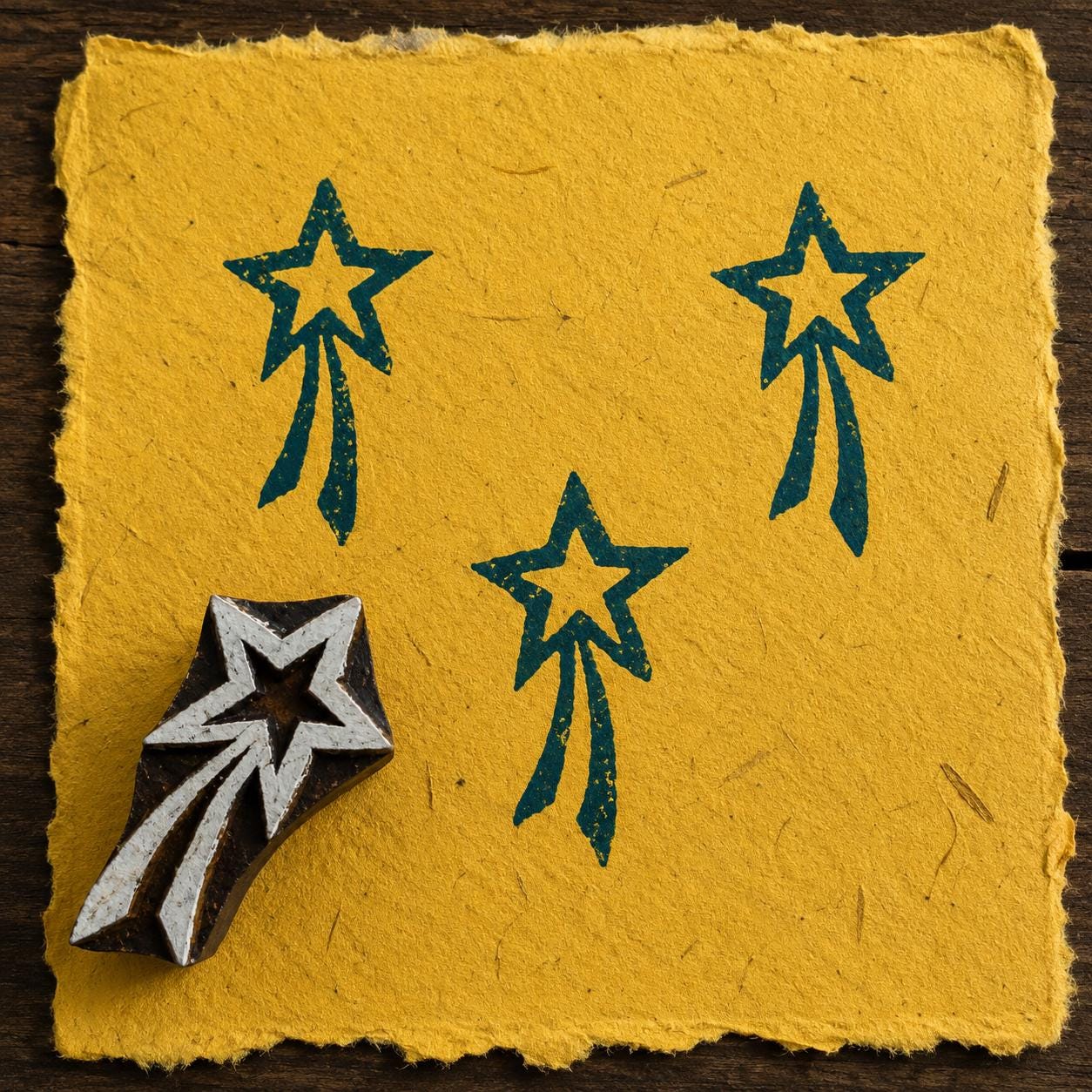 Hand-carved wooden shooting star printing block with teal blue celestial impressions on textured mustard handmade paper.