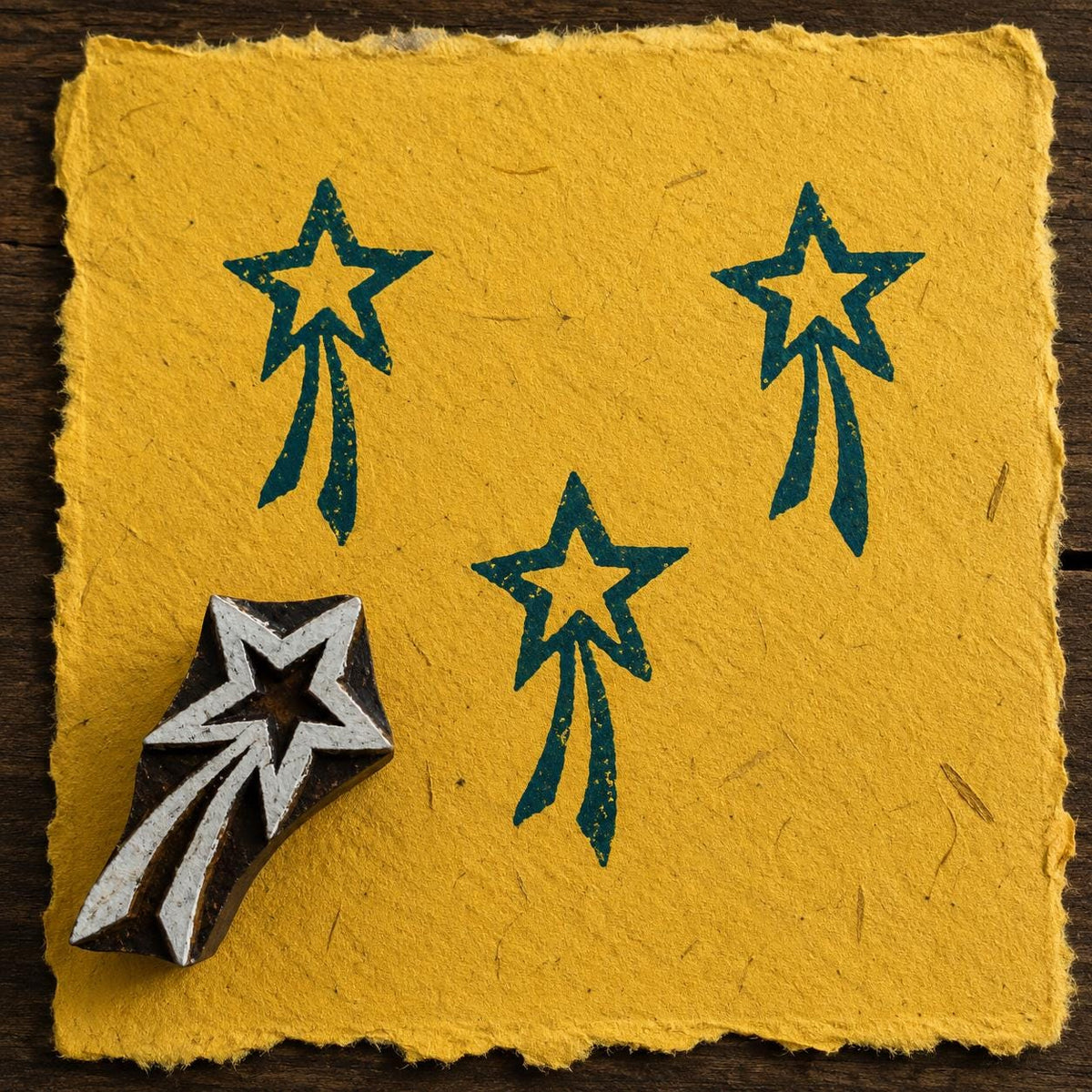 Hand-carved wooden shooting star printing block with teal blue celestial impressions on textured mustard handmade paper.