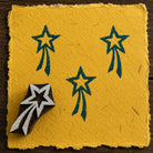 Hand-carved wooden shooting star printing block with teal blue celestial impressions on textured mustard handmade paper.