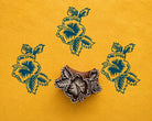 Hand carved Indian wood printing block featuring a detailed hibiscus flower with leaves, shown with three blue floral impressions on textured yellow handmade paper.