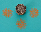 Hand carved Indian textile printing block with a radial teardrop star pattern, shown with three orange impressions on turquoise handmade paper.