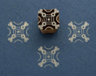Hand carved wooden printing block with a symmetrical geometric Cornerstone Pattern, shown with pale impressions on textured blue handmade paper.