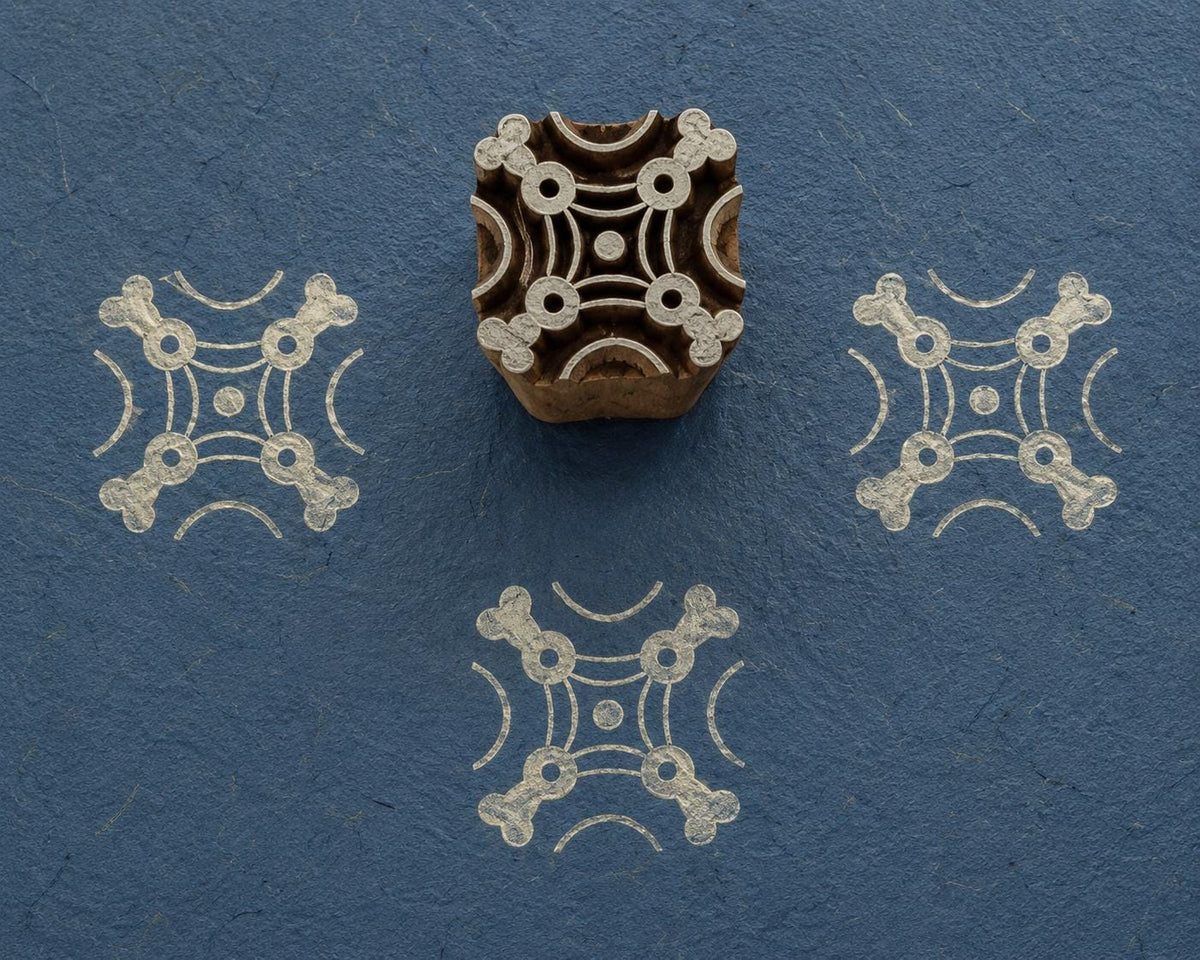Hand carved wooden printing block with a symmetrical geometric Cornerstone Pattern, shown with pale impressions on textured blue handmade paper.