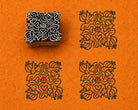 Hand carved wooden printing block with a dense folk floral motif of flowers, curling leaves, fruit-like forms and a central heart, shown with three blue impressions on textured orange handmade paper.