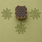 Hand carved wooden printing block with an intricate repeating swirl motif, shown with three dark green impressions on textured sage green handmade paper.