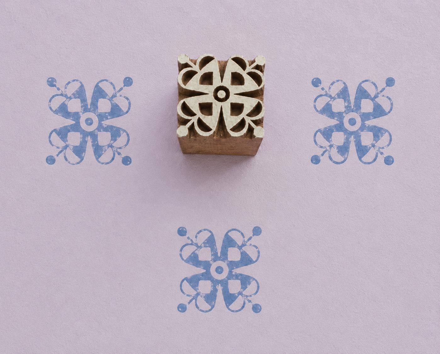 Hand carved wooden printing block featuring a symmetrical Ribbon Bloom motif with looping petals and rounded details, shown with three blue impressions on lavender handmade paper.