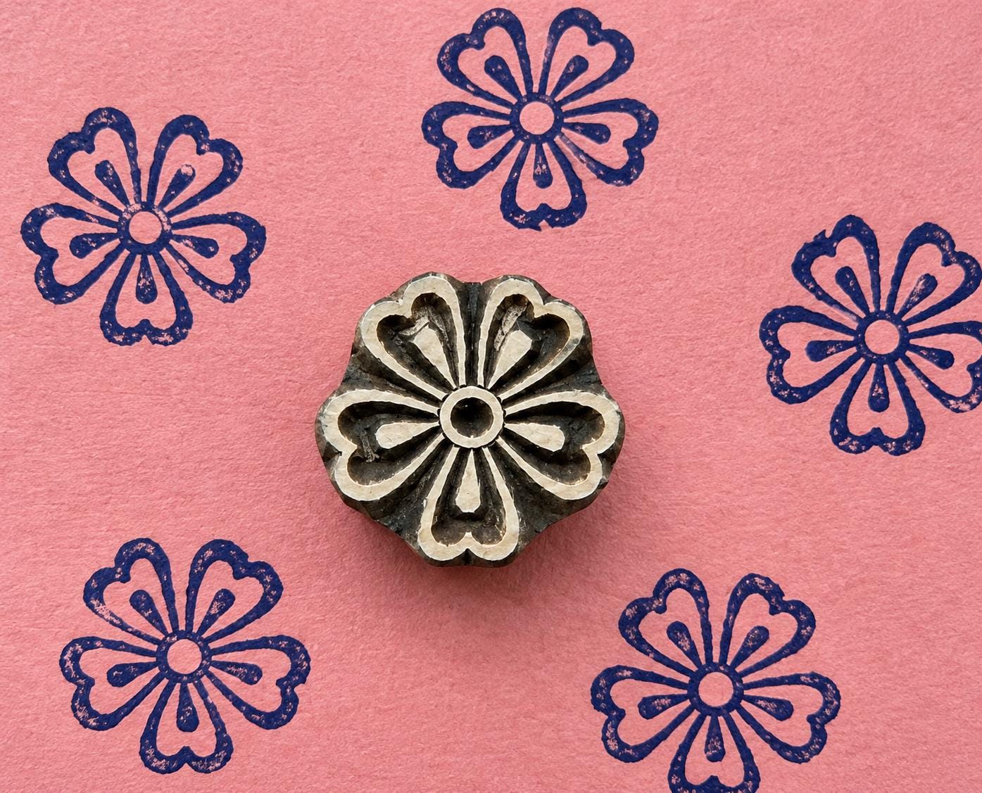 Hand carved wooden printing block with six heart-shaped petals around a circular centre, shown with five navy floral impressions on textured pink handmade paper.
