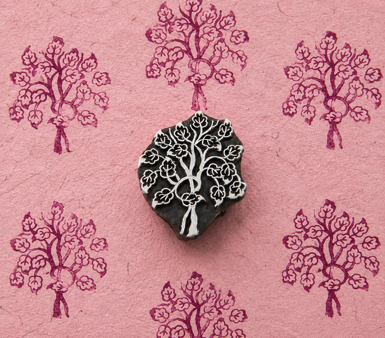 Hand carved wooden printing block featuring a flowering tree motif, surrounded by repeated purple blossom tree impressions on textured pink handmade paper.
