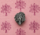 Hand carved wooden printing block featuring a flowering tree motif, surrounded by repeated purple blossom tree impressions on textured pink handmade paper.