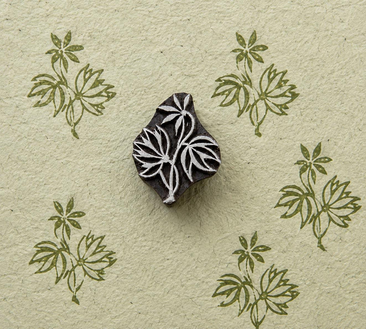 Hand-carved wooden printing block featuring a three-leaf botanical sprig with a small flower, shown with repeated green impressions on textured handmade paper.