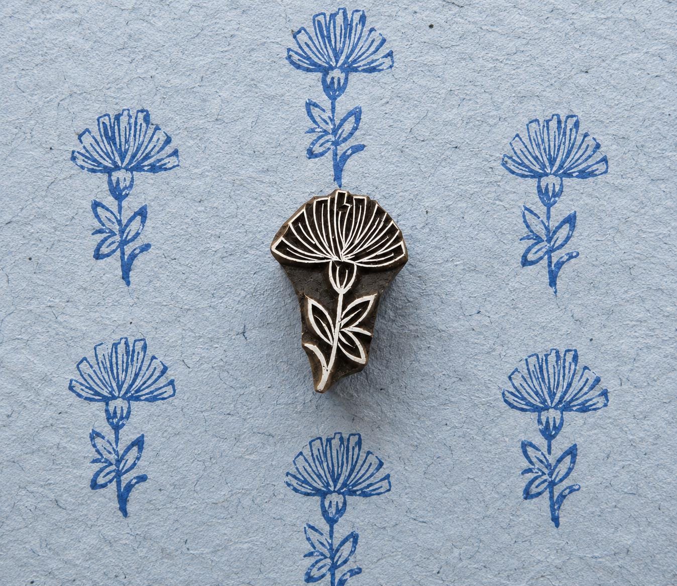 Hand-carved wooden printing block featuring a tall thistle-like flower with a finely detailed bloom, stem and leaves, shown with repeated blue impressions on textured handmade paper.