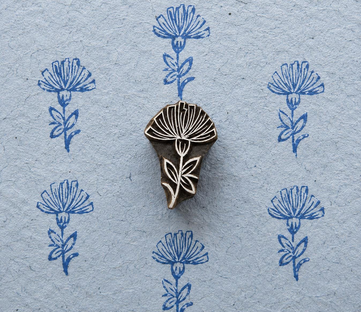 Hand-carved wooden printing block featuring a tall thistle-like flower with a finely detailed bloom, stem and leaves, shown with repeated blue impressions on textured handmade paper.