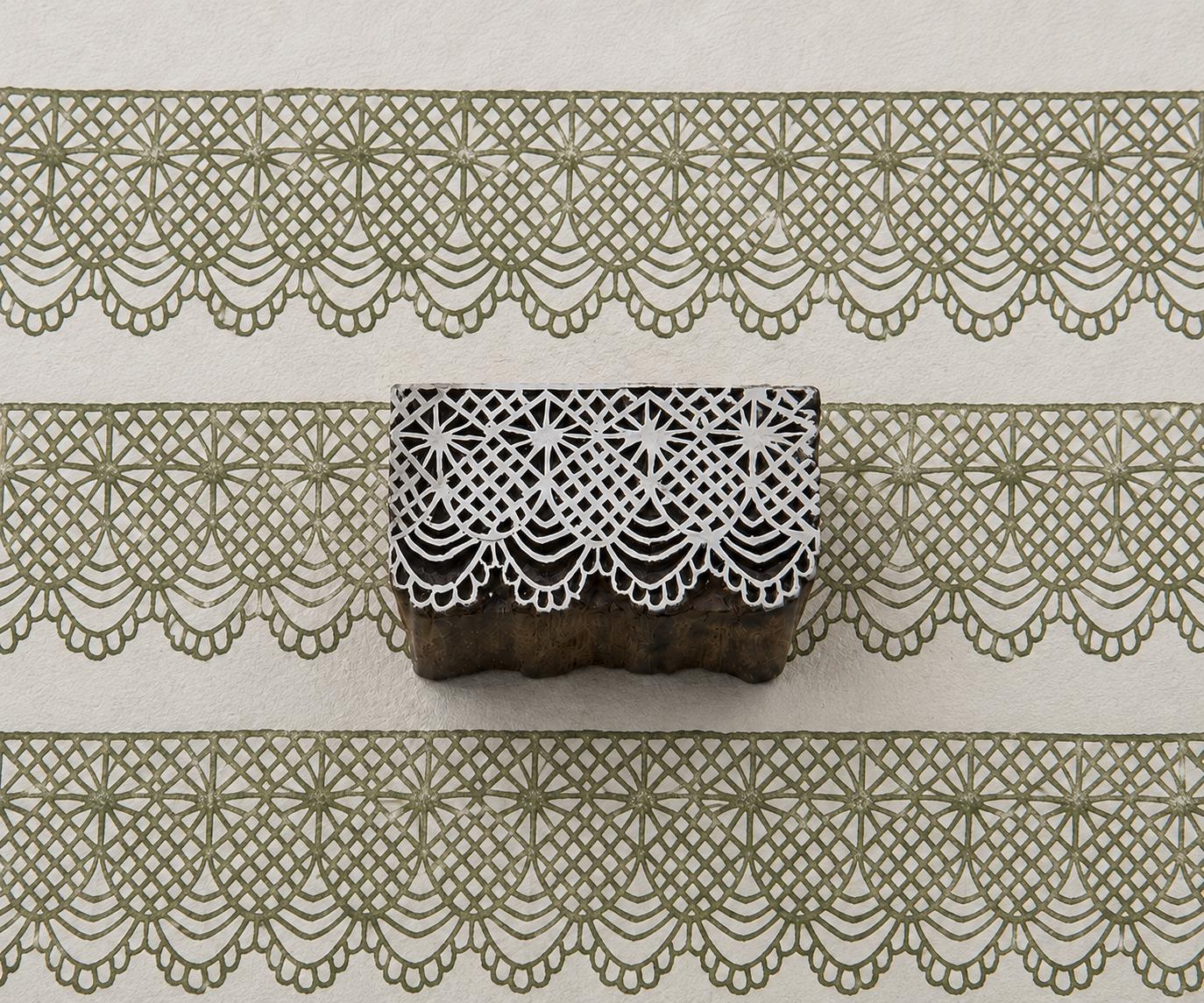 Hand-carved wooden printing block with an intricate diamond lattice and scalloped ornamental border, shown with repeated green impressions on textured handmade paper.