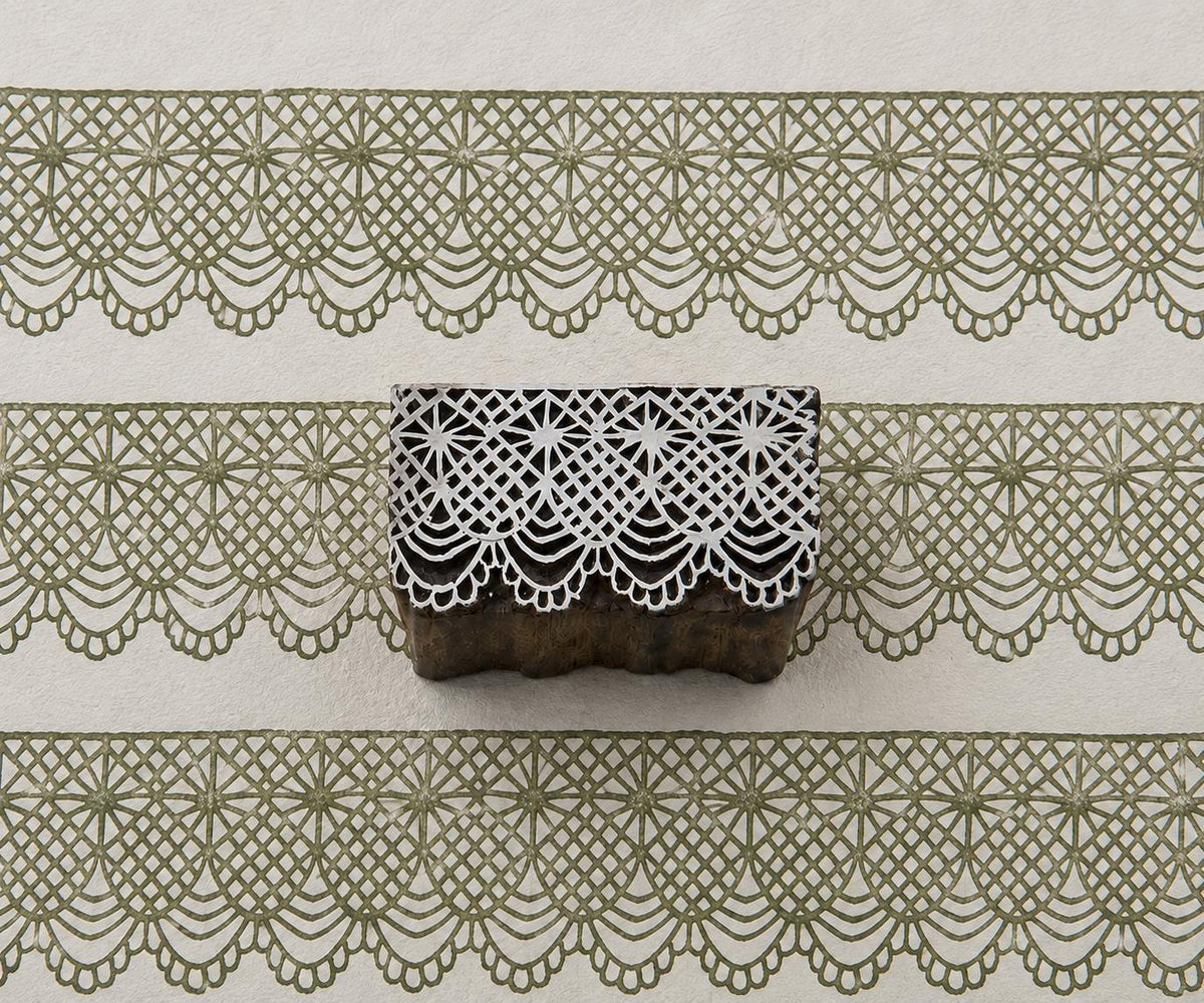Hand-carved wooden printing block with an intricate diamond lattice and scalloped ornamental border, shown with repeated green impressions on textured handmade paper.