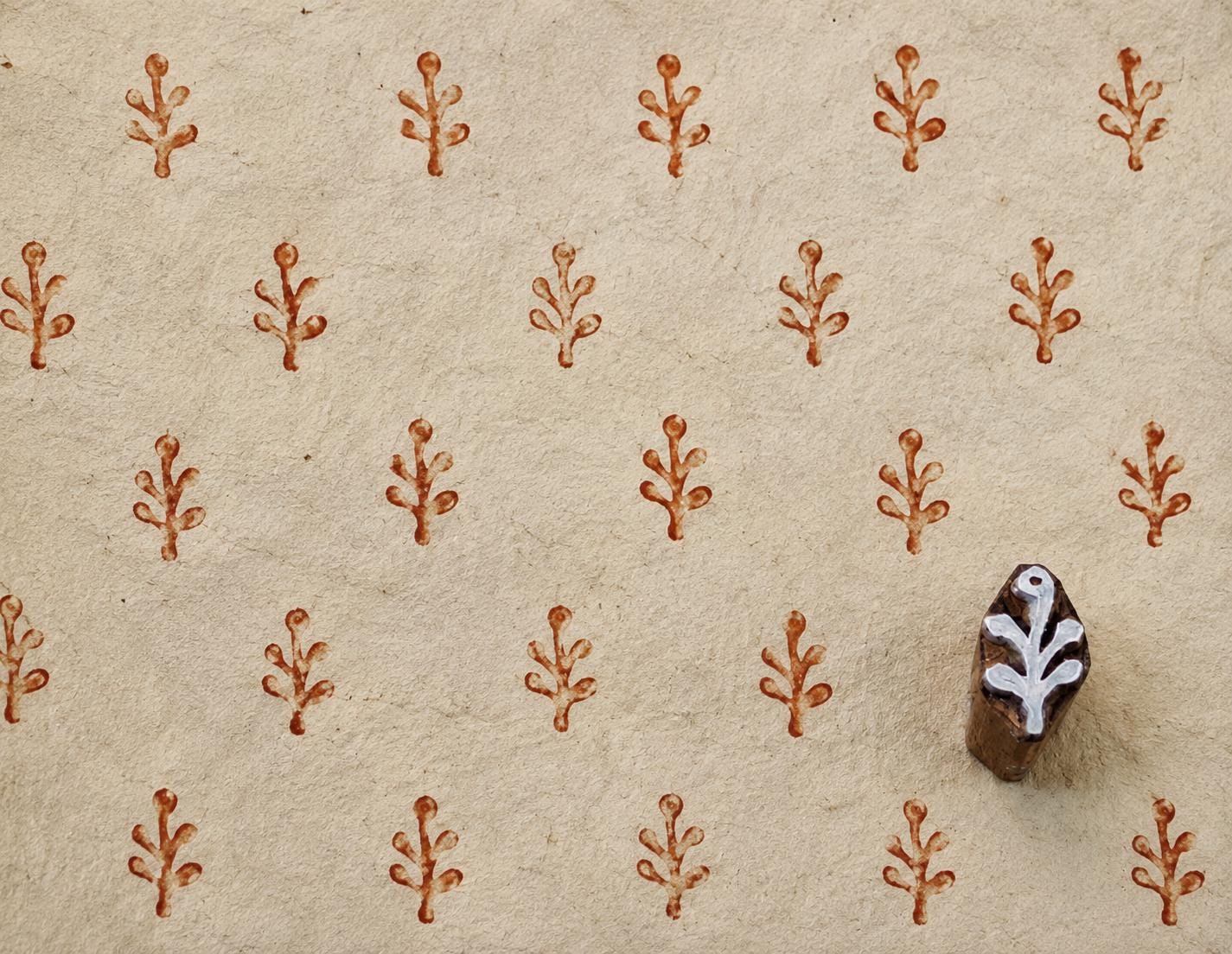 Hand-carved 2 cm wooden printing block with a small branch motif, shown beside repeated reddish-brown impressions on handmade cream paper.