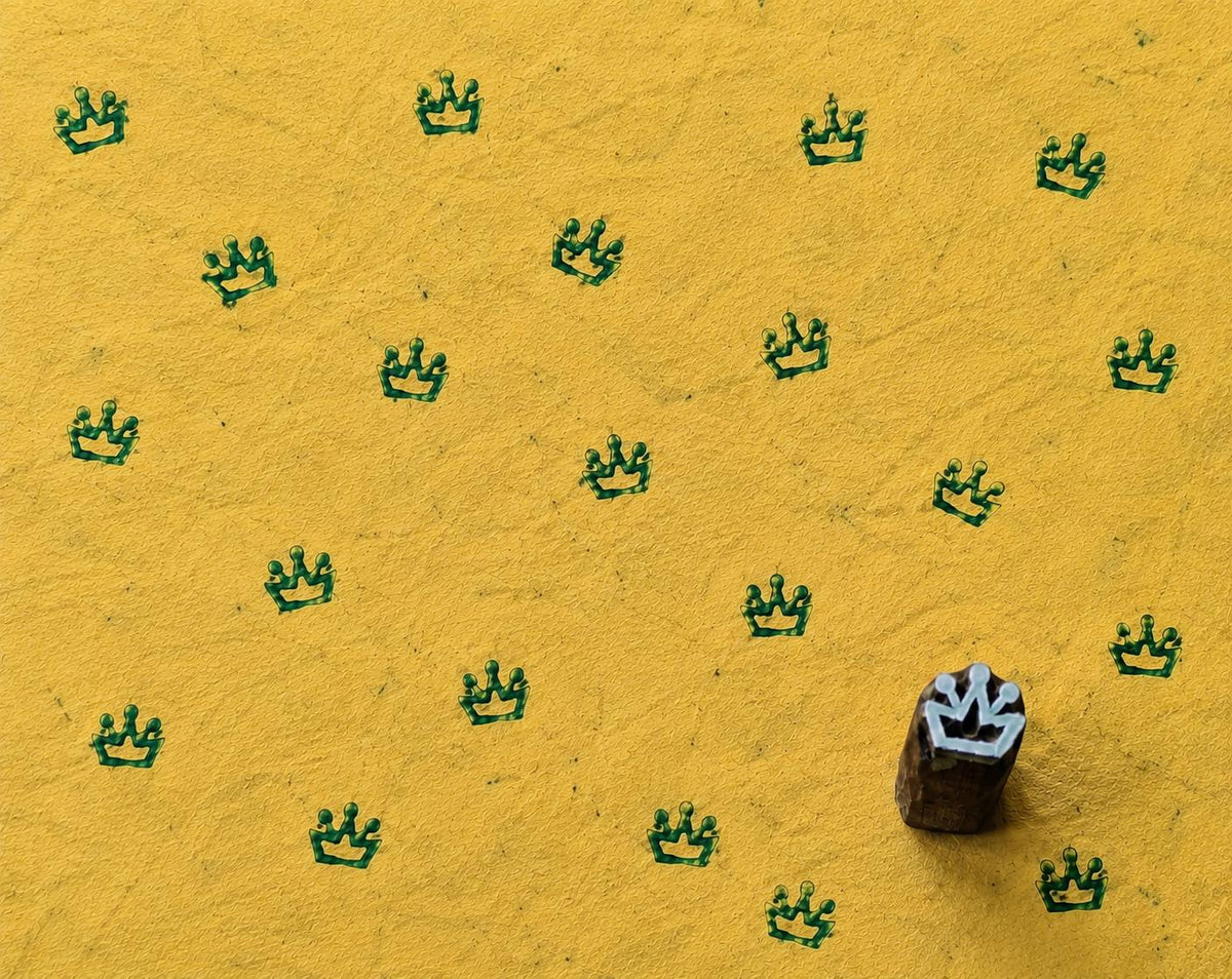 Small hand-carved wooden printing block with a crown motif beside repeated green crown impressions on handmade yellow paper.
