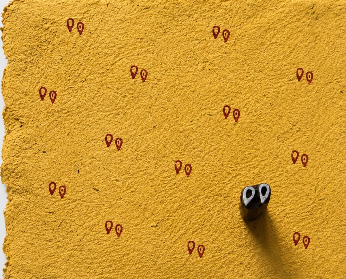 Hand carved wooden printing block with a paired teardrop motif, shown beside repeated red impressions on textured handmade yellow paper.
