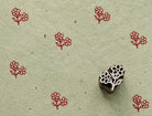 Hand carved wooden printing block measuring approximately 2.8 × 2.5 cm, featuring two rounded blossoms on a branching stem, shown with repeated red impressions on textured handmade paper.