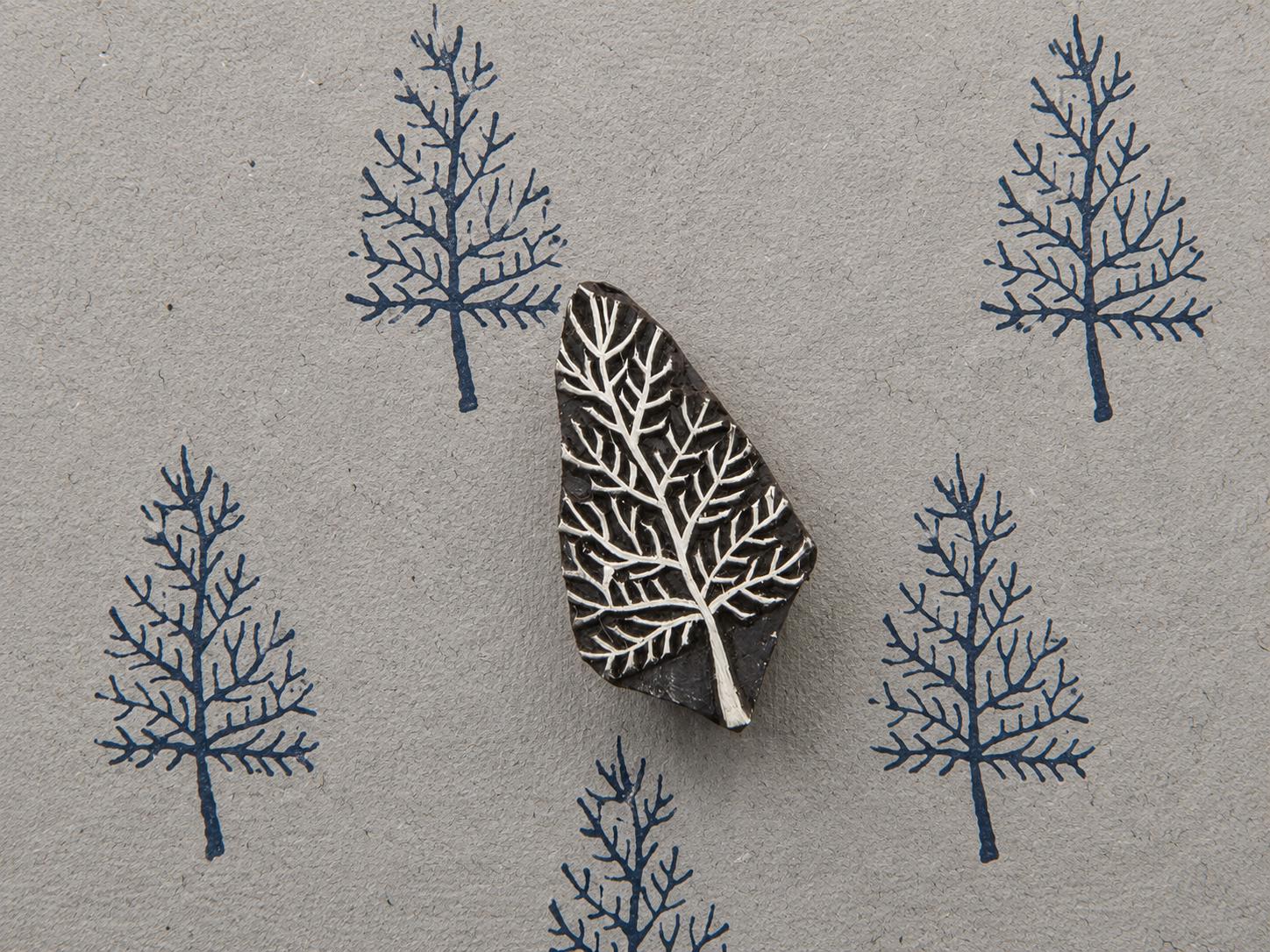 Hand carved bare tree wooden block stamp displayed on textured handmade paper with repeated blue tree impressions.