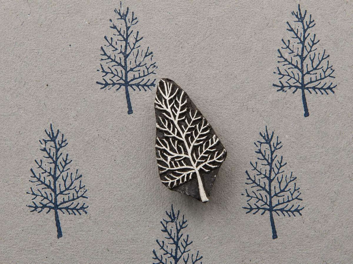 Hand carved bare tree wooden block stamp displayed on textured handmade paper with repeated blue tree impressions.