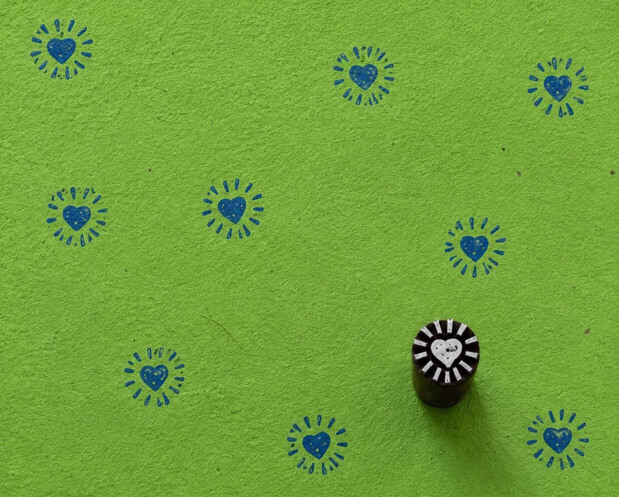 Hand carved heart sunburst wood printing block shown on green handmade paper with repeated blue heart prints
