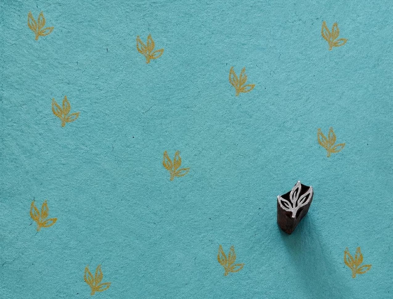 Hand-carved wooden three-leaf sprig printing block beside repeated ochre leaf impressions on textured turquoise handmade paper.