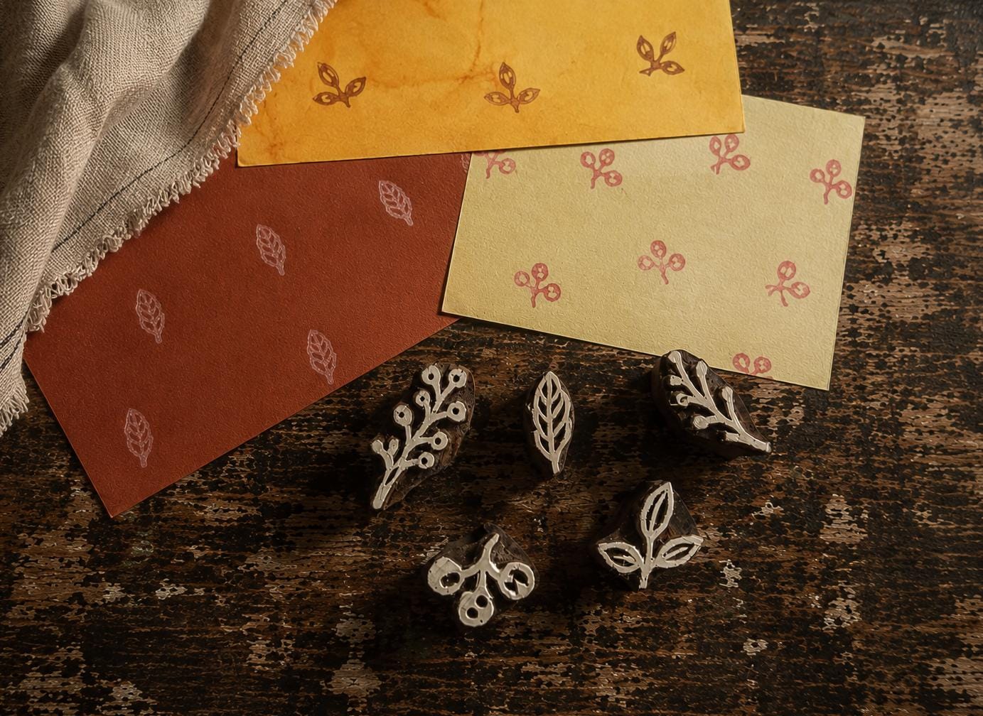 Set of five tiny hand carved wooden botanical printing blocks displayed on a distressed wooden surface with handmade paper samples showing their printed designs.