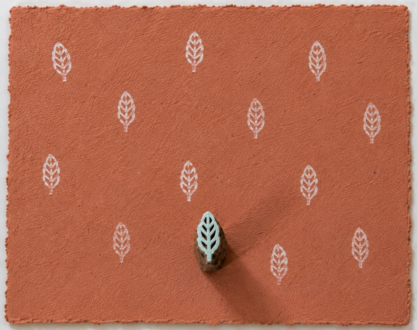 Tiny hand carved wooden leaf printing block, shown with repeated white leaf impressions on terracotta handmade paper.