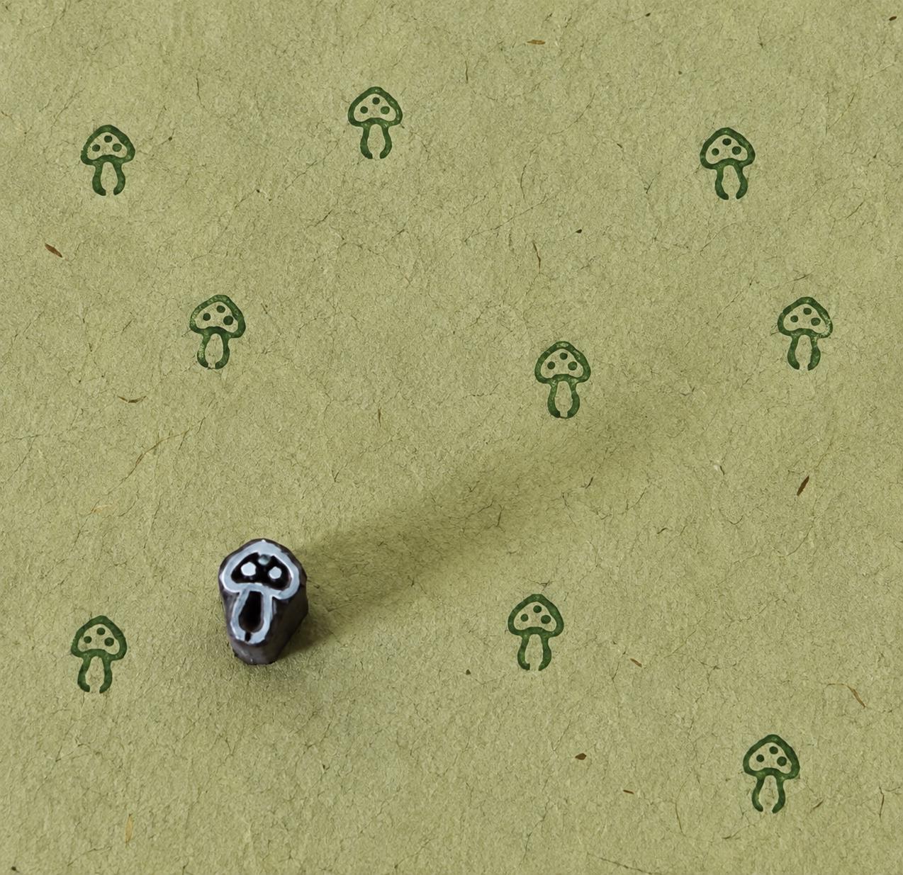 Tiny hand carved wooden mushroom printing block shown with repeated green toadstool impressions on textured handmade paper.