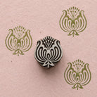 Hand carved wooden ornamental lotus stamp shown with repeated olive green floral impressions on textured pink handmade paper.