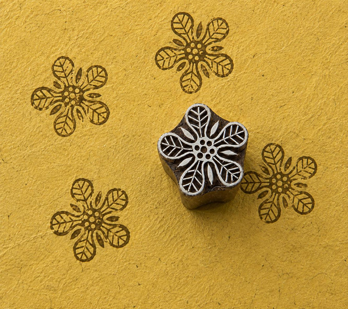 Hand-carved wooden flower stamp with five leafy petals, shown with brown impressions on textured mustard handmade paper.