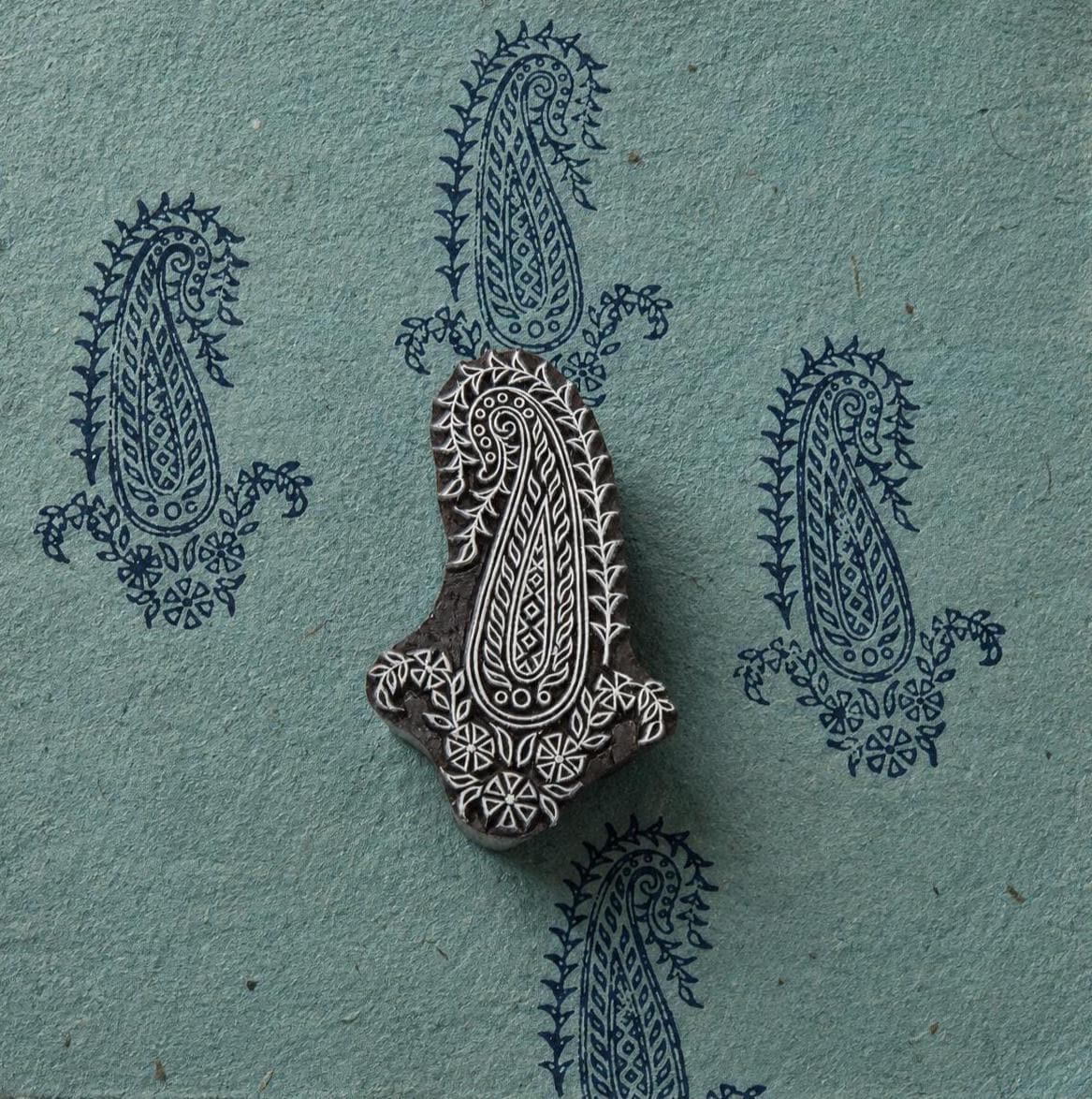 Hand carved Indian wooden printing block featuring an intricate jewel paisley motif with floral and geometric details, shown with repeated navy impressions on handmade paper.