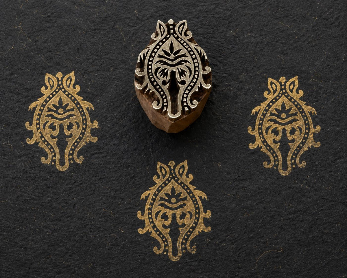 Hand carved wooden printing block featuring a symmetrical Temple Ornament motif with scrolling details, dotted borders and a stylised floral centre, shown with three gold impressions on dark handmade paper.