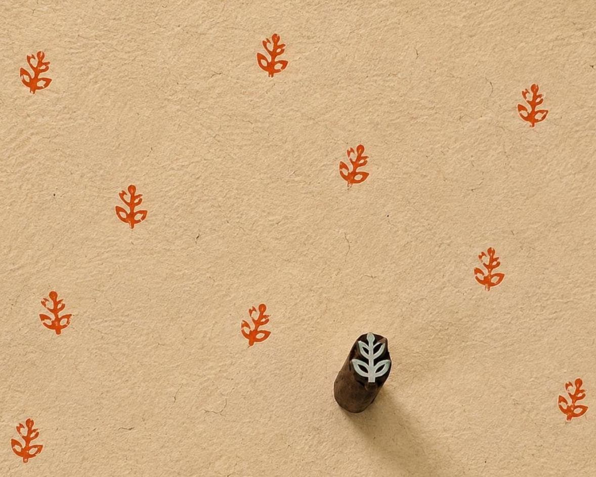 Hand carved Indian wooden printing block with a small stylized leaf motif, shown creating a repeating pattern on handmade paper.