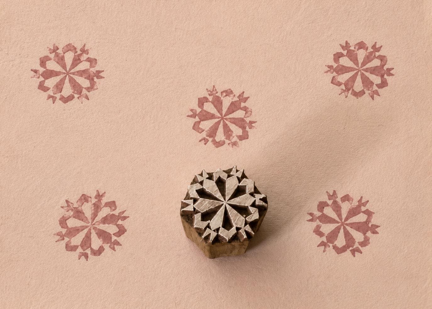 Hand-carved wooden printing block featuring a symmetrical folk starburst motif, shown with five dusty rose impressions on textured handmade paper.