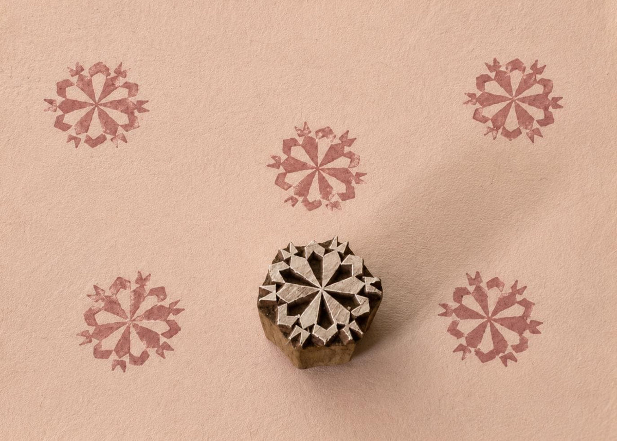 Hand-carved wooden printing block featuring a symmetrical folk starburst motif, shown with five dusty rose impressions on textured handmade paper.