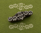 Hand-carved wooden printing block with symmetrical floral scrollwork and curling botanical motifs, shown with white printed impressions on green handmade paper.